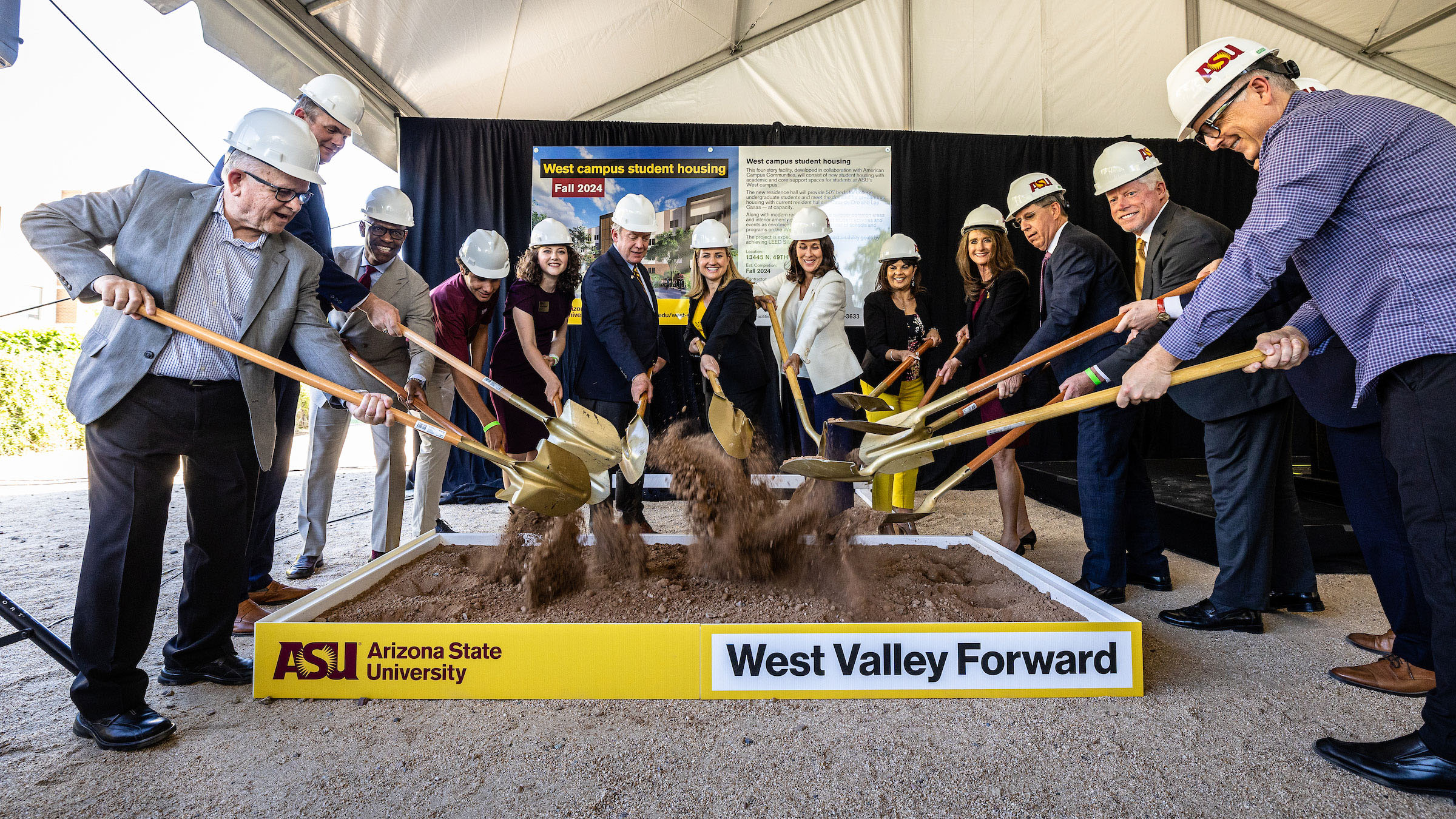 A group of 13 men and women, made up of community, political and ASU leaders, all dressed in business clothing and hard hats, use gold shovels to perform a ceremonial groundbreaking at the ASU West Campus.