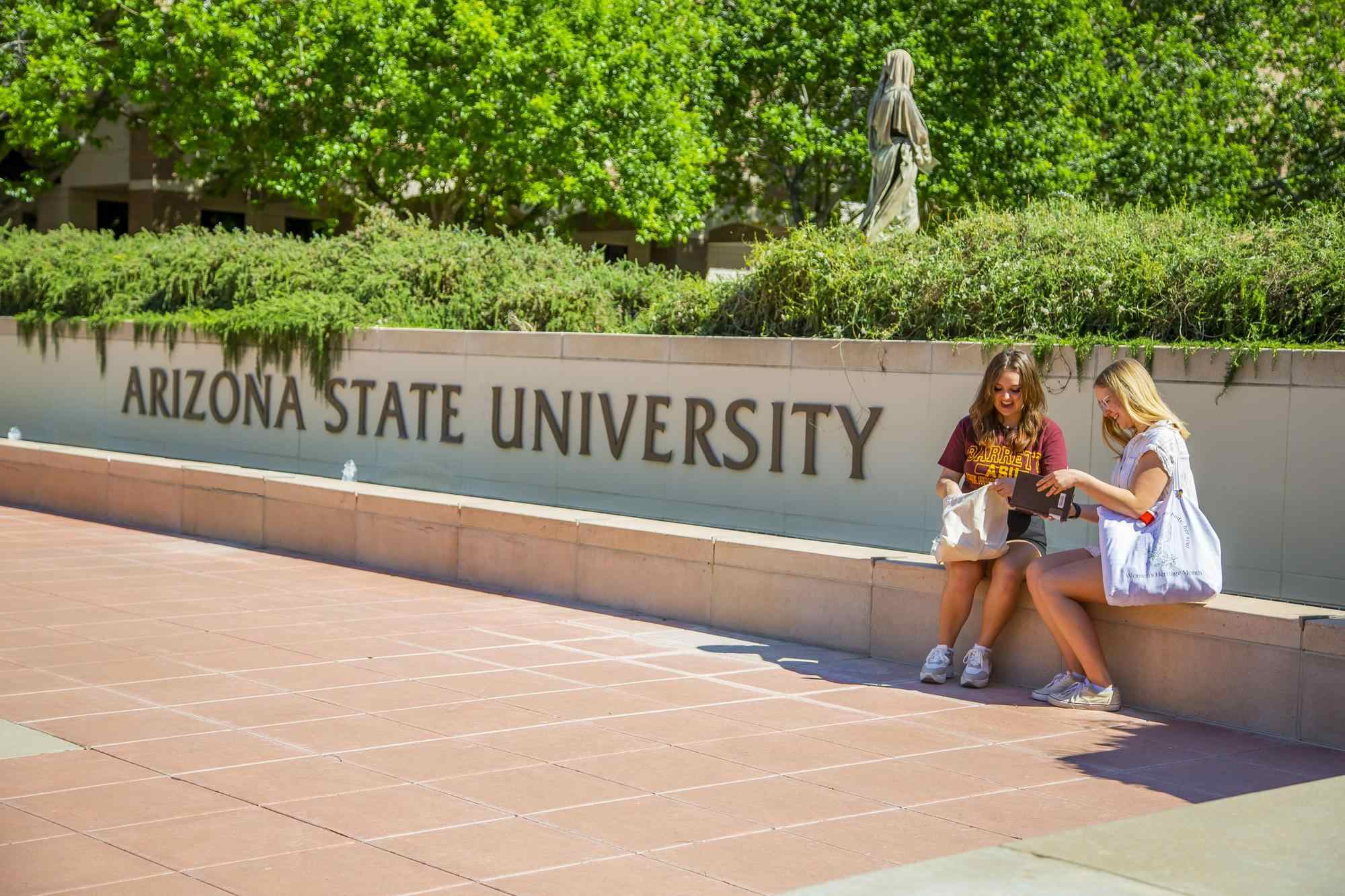 Two students sit near the large "Arizona State University" sign on the ASU West Valley campus